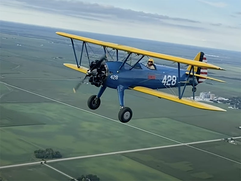 vintage biplane with blue fuselage and yellow wings flying above farmland